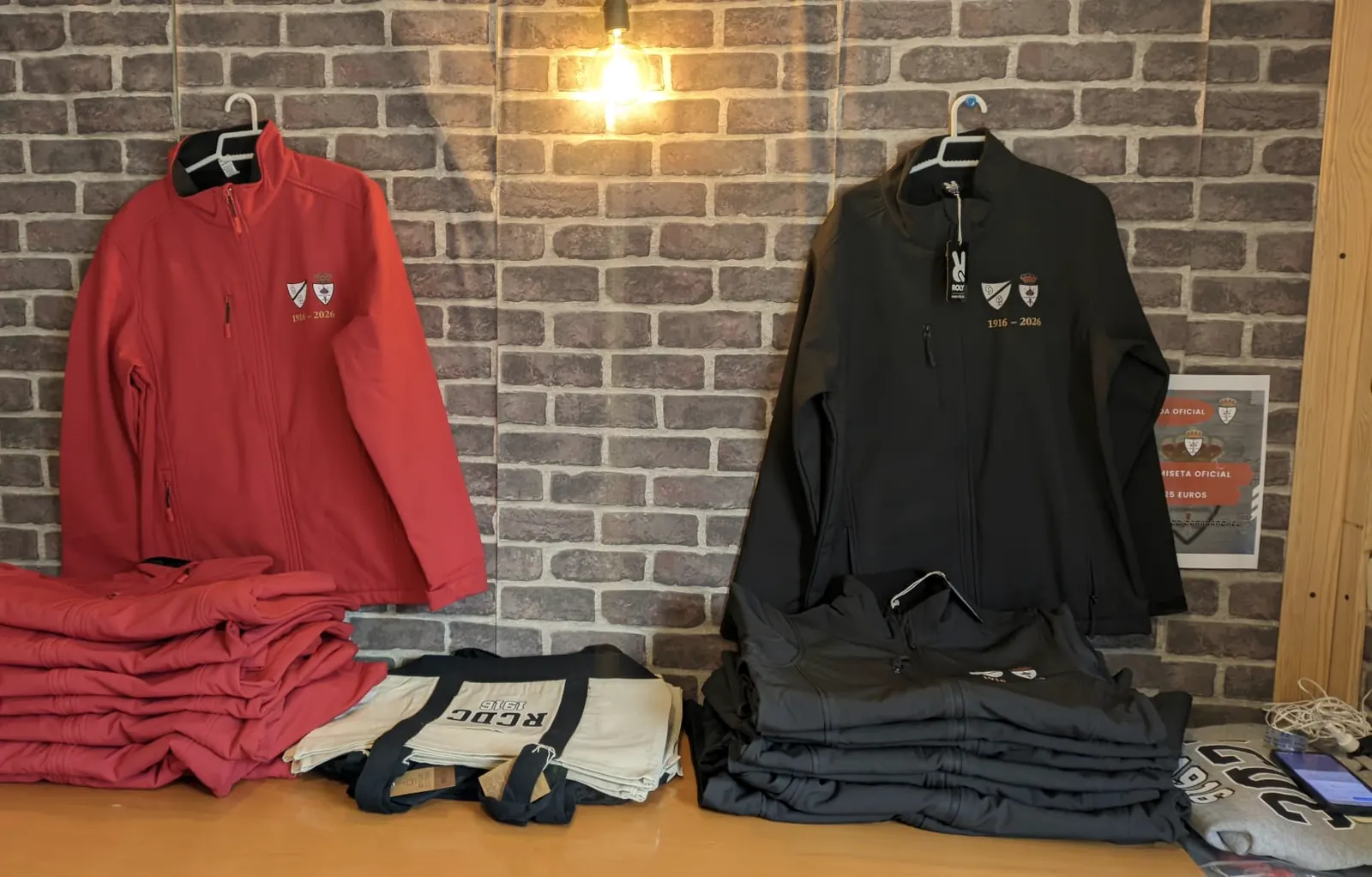 Cortavientos rojo y negro del 110 aniversario y bolsas RCDC 1916 en la tienda del club